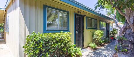 A single-story house with a yellow facade and blue trim, a black door, and a small front yard with various plants.