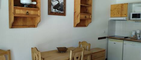 Une table de salle à manger en bois avec des chaises, un four à micro-ondes blanc et un lave-vaisselle blanc dans une cuisine dotée de meubles en bois et d’un tableau encadré au mur.