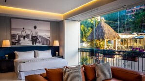 Una habitación de hotel con cama, sofá, un ventanal con vistas a una construcción de techo de paja y un cuadro en la pared.