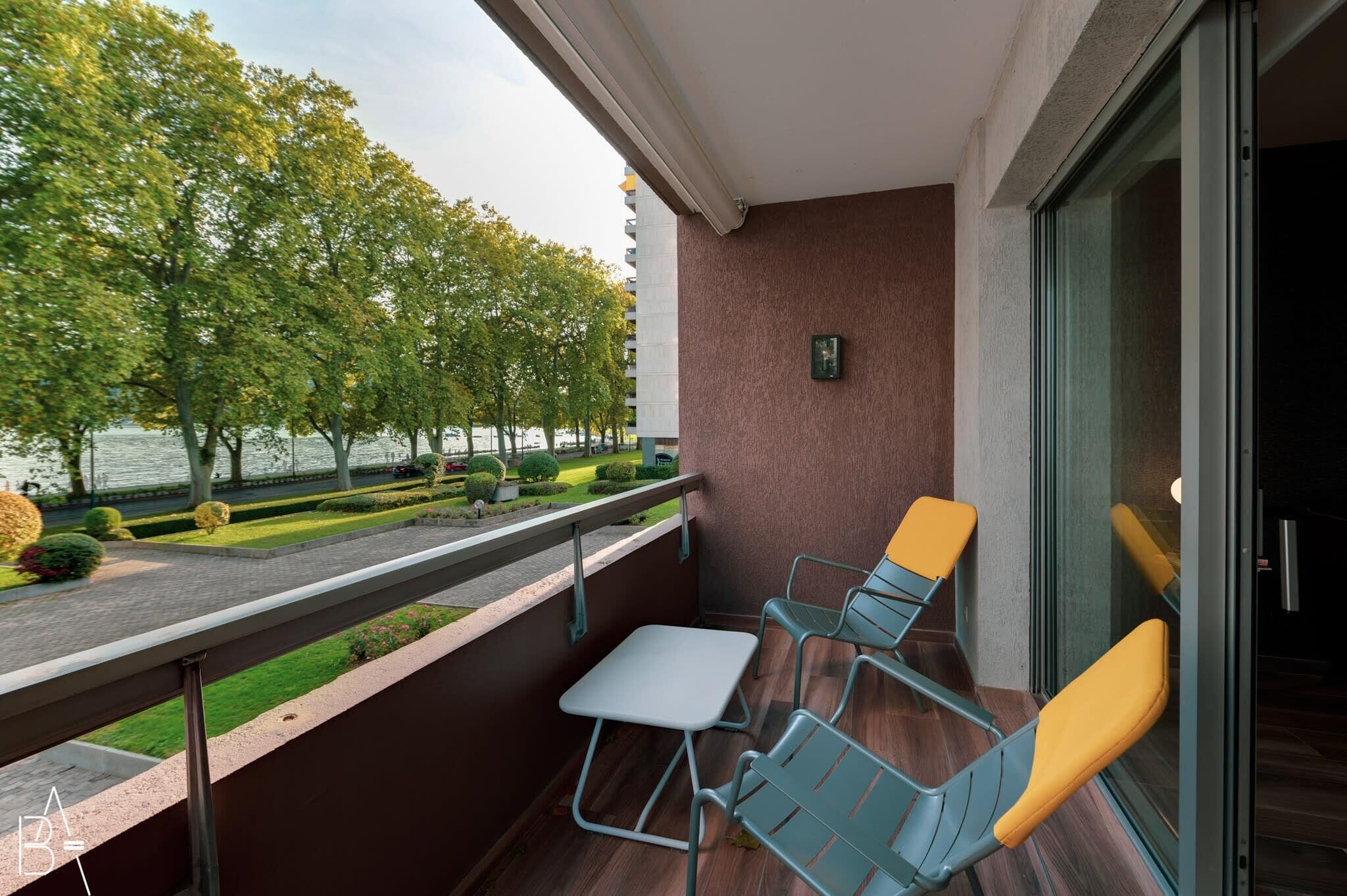A balcony with a table and two chairs, overlooking a landscaped area with trees and a body of water.