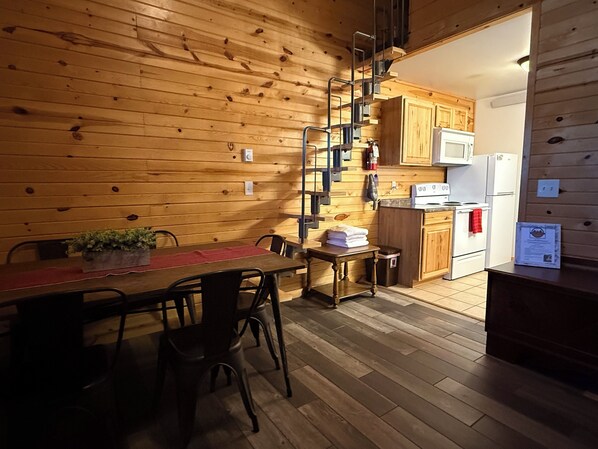 A wooden interior with a dining table, chairs, a kitchen area with cabinets, a microwave, and a refrigerator. - Frenchy's Hideaways Cabin #6 (Custer)