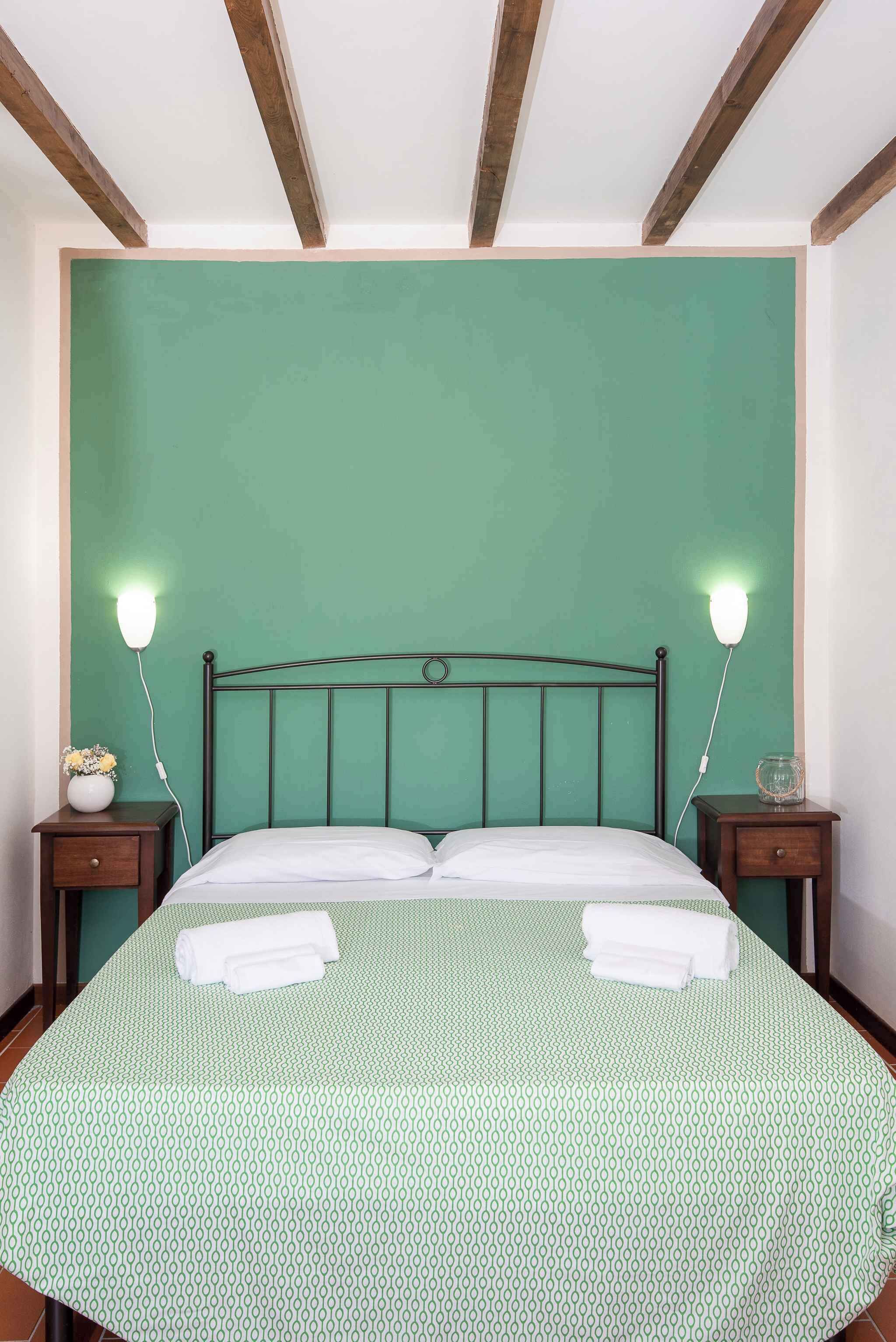 Una cama bien tendida con una colcha verde estampada, dos lámparas de noche y una mesita de noche con un florero.