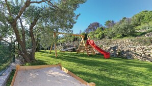 Une aire de jeux avec un toboggan et des balançoires, entourée d’une pelouse et d’un mur en pierre.