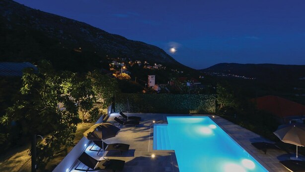 Une vue nocturne d’une piscine, avec des chaises longues et des parasols, entourée de verdure et bordée d’une colline où se trouvent des maisons.