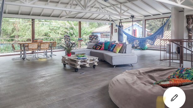 A spacious living area with a white sofa, a dining table with chairs, a hammock, and a view of the surrounding greenery.