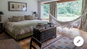 A bedroom with a bed, a bedside table with books, a hammock, and a view of the outdoors.