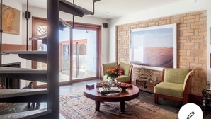 A living room with a brick wall, a large window, a green armchair, a wooden coffee table, and a staircase.
