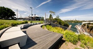 A curved wooden boardwalk with a concrete edge, leading to a waterfront area with a view of boats and a city skyline in the distance.