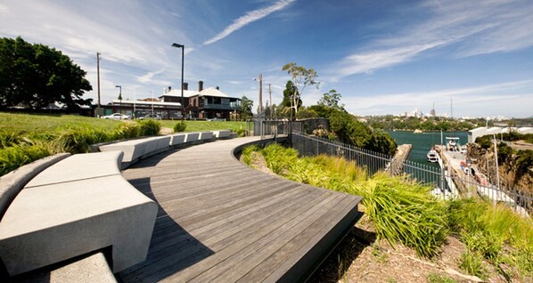 A curved wooden boardwalk with a concrete edge, leading to a waterfront area with a view of boats and a city skyline in the distance.