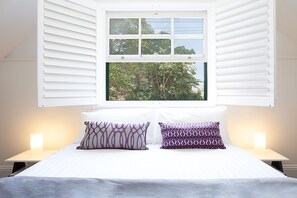 A bedroom with a bed, two bedside lamps, a window with shutters, and a view of trees.