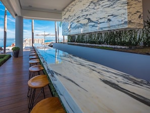Un bar junto a la piscina con mostrador de mármol y taburetes de madera, con vista a una masa de agua y un paisaje urbano al fondo.
