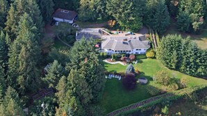 An aerial view of a large, multi-story house surrounded by dense forest, with a well-maintained lawn and a driveway leading to the entrance.