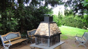 A rustic outdoor seating area with a stone-built wood stove, surrounded by benches and lush greenery.
