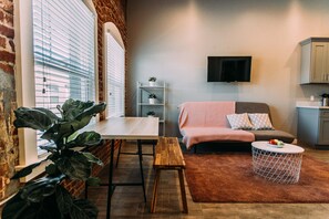 Uma sala de estar moderna com parede de tijolos, uma mesa branca com um banco, sofá cinza com manta rosa, mesa de centro redonda e televisão fixada na parede.