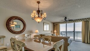 Habitació d'hotel amb taula de menjador, cadires, un penjoll, un mirall gran, un sofà, un balcó amb vista a l'oceà i un ventilador de sostre.