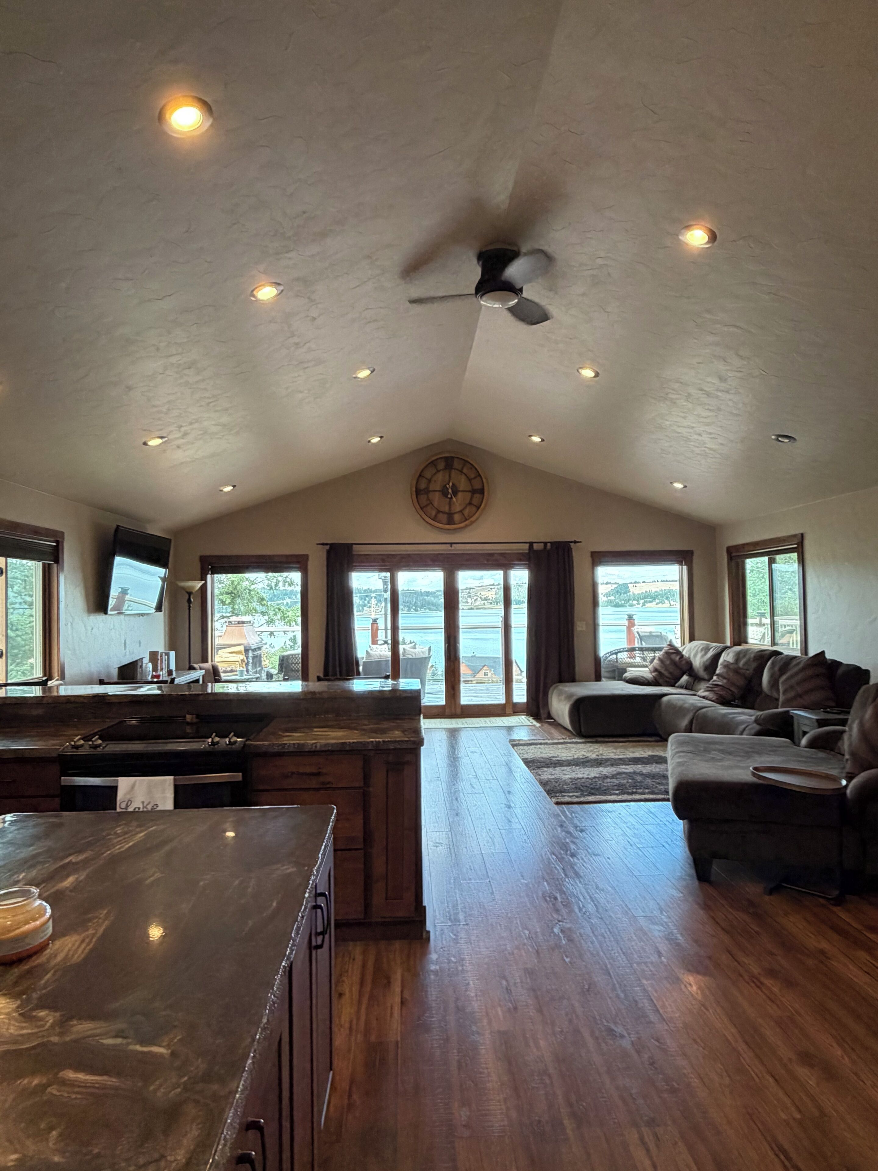 A spacious living area with a kitchen, large windows, and a ceiling fan.