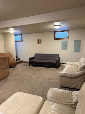 A room with a brown leather sofa, a beige leather couch, and a brown leather chair. There are two windows with blinds, a wall-mounted light fixture, and a small framed picture.