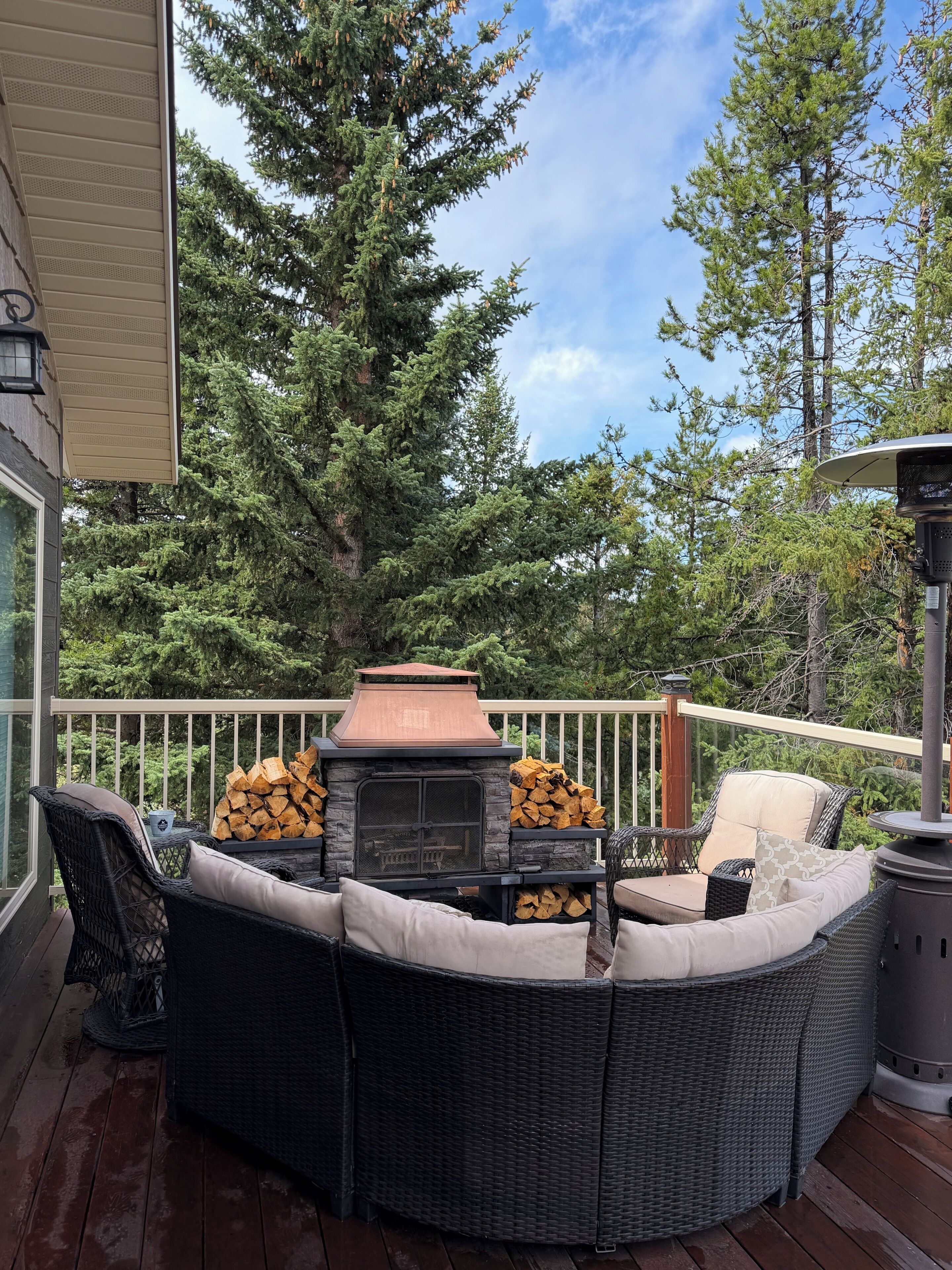 An outdoor deck with a stone fireplace, wicker furniture, and a view of surrounding trees.