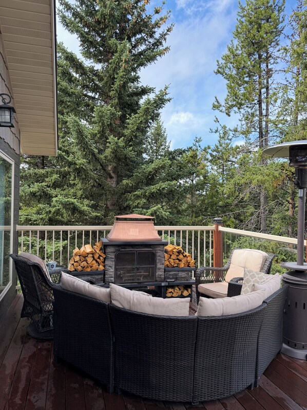 An outdoor deck with a stone fireplace, wicker furniture, and a view of surrounding trees.