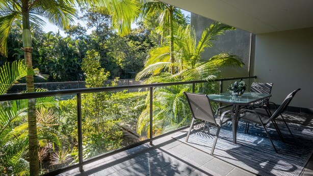 Een balkon met een glazen tafel en stoelen, met uitzicht op een weelderige tuin vol planten en bomen.