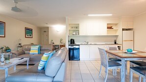 Een moderne woonkamer met een bank, een eettafel met stoelen, een keuken met kasten en apparatuur, en een plafondventilator.