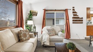 Een woonkamer met een beige bank, een bijzettafeltje en een potplant. Er is een raam met jaloezieën en een bord op de muur.