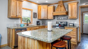 Una cocina con gabinetes de madera, encimeras de granito, una isla central con taburetes, una estufa y un letrero que dice "let's eat".