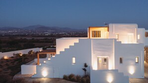 A modern white building with multiple levels and illuminated windows, surrounded by a landscaped area with sparse vegetation.
