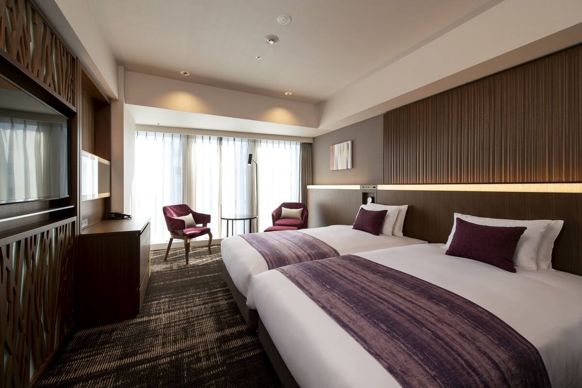 Quarto de hotel com duas camas, televisão, escrivaninha, cadeira e vista para o exterior.