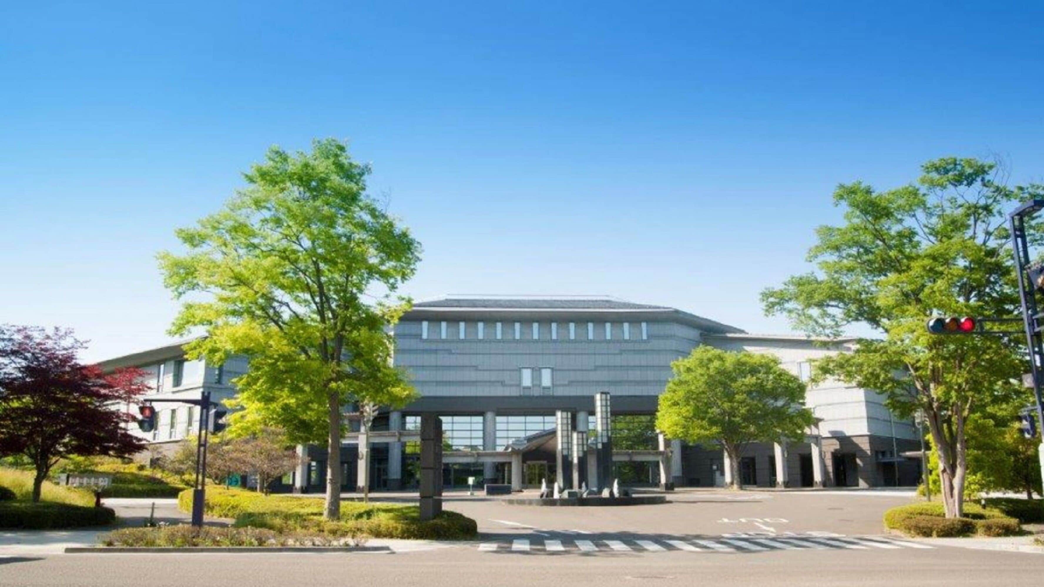 A modern building with a flat roof and large windows, surrounded by trees and a clear sky.