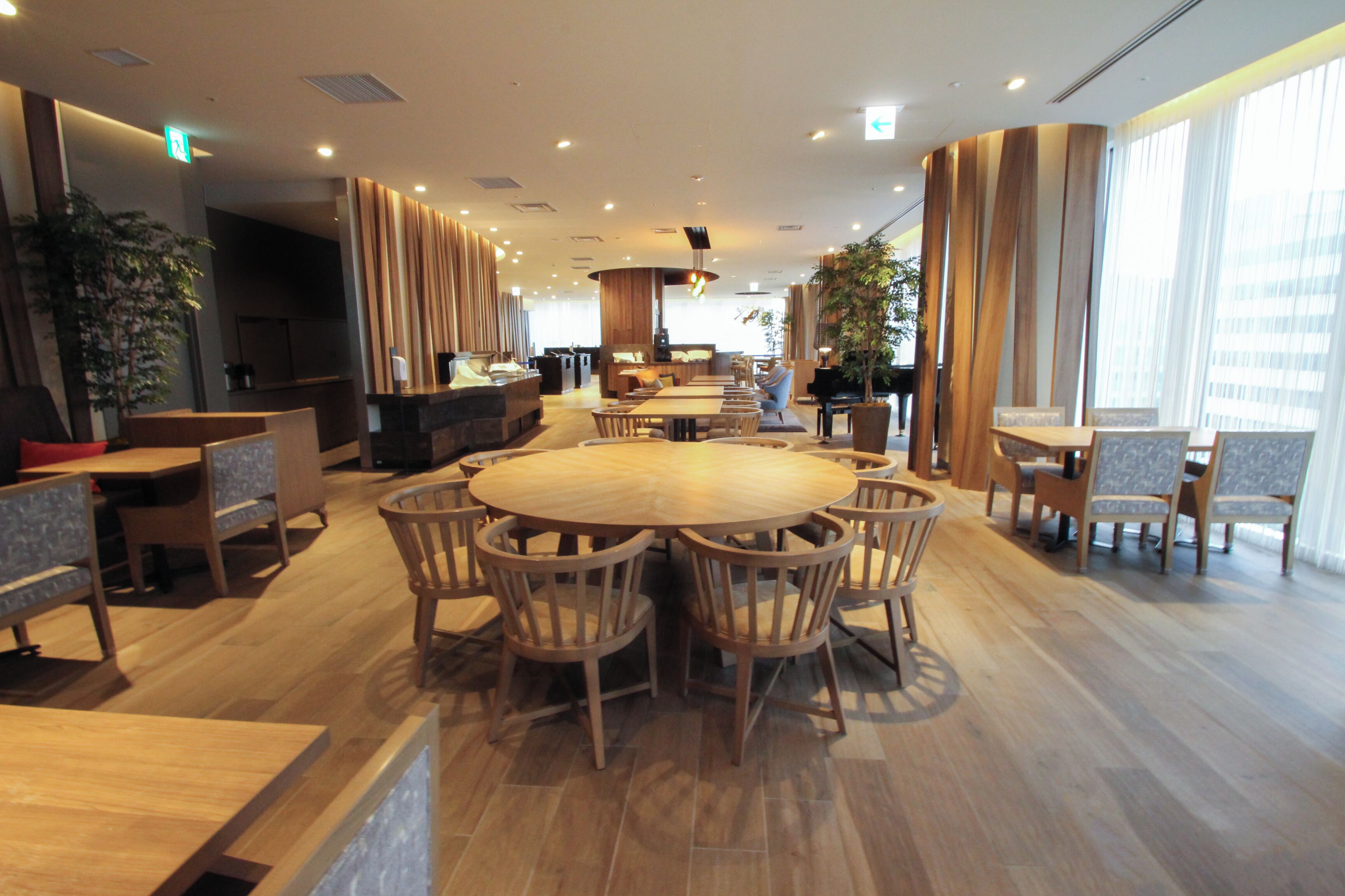 A spacious hotel lobby with a central round wooden table surrounded by chairs, a reception desk, and a seating area with sofas.
