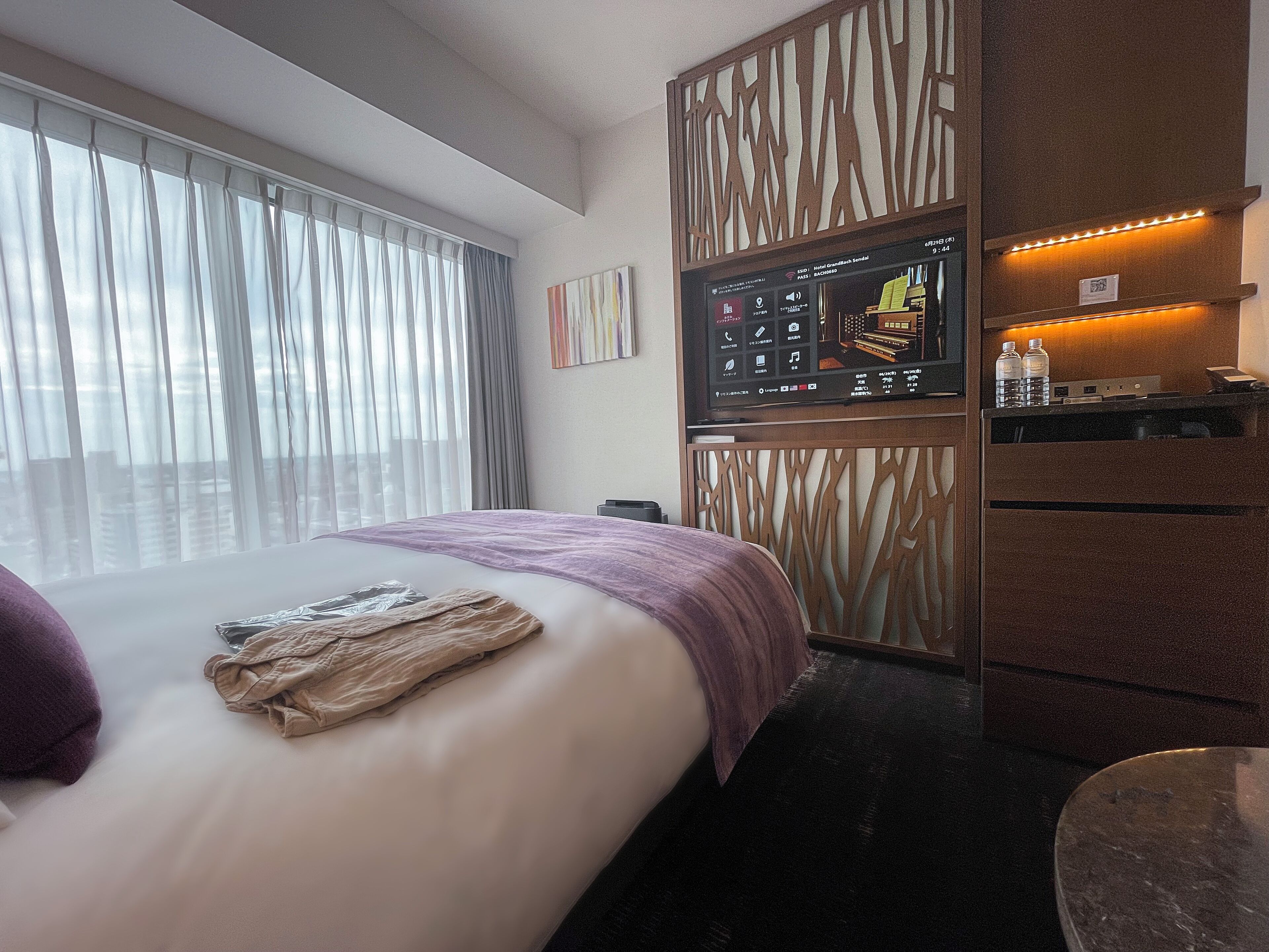Ein Hotelzimmer mit einem Bett, einem an einer Holzplatte montierten Fernseher, einem Schreibtisch mit Stuhl und einem Fenster mit Vorhängen.