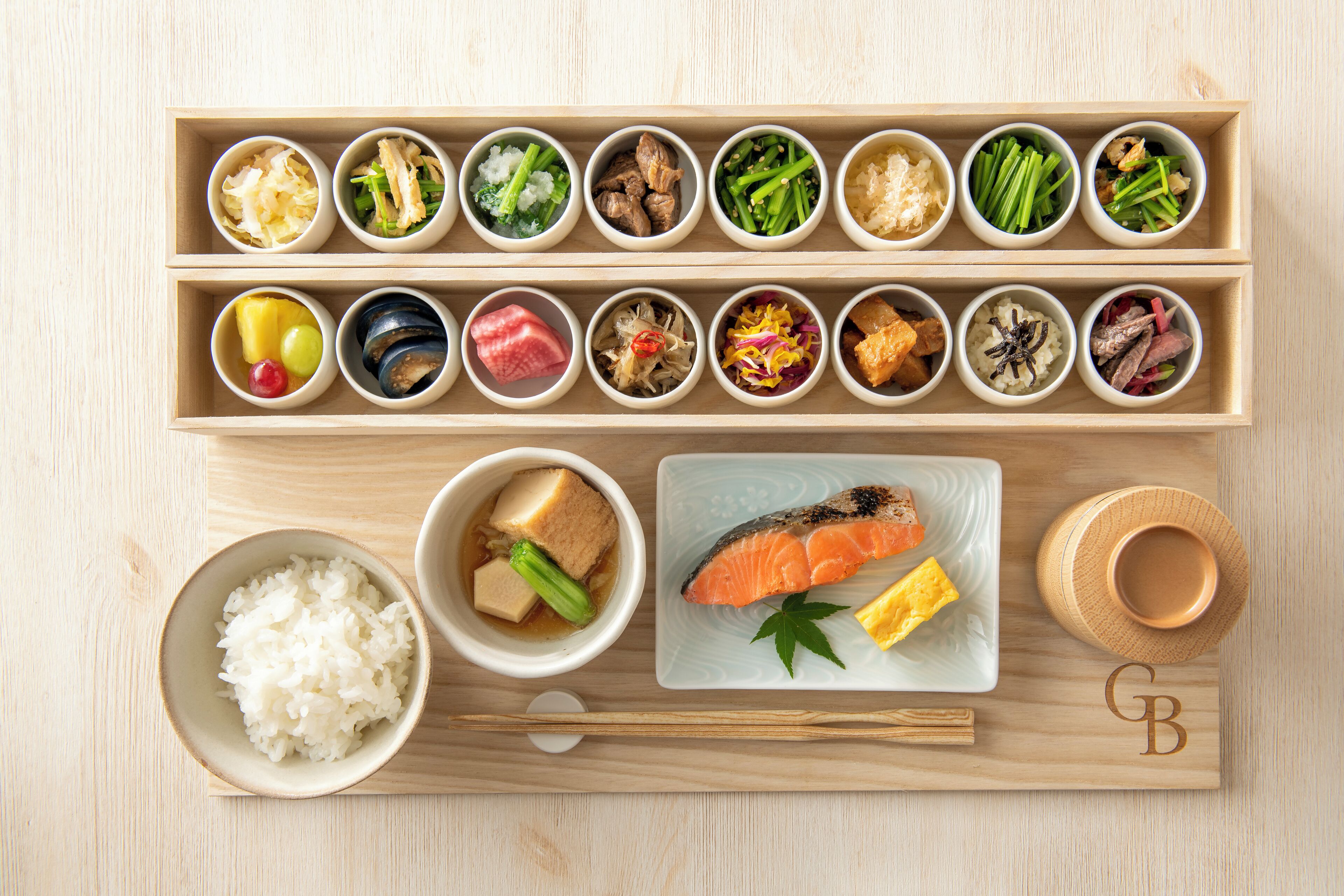 A wooden tray with multiple small bowls containing various food items, a bowl of rice, a plate with salmon and egg, and chopsticks.