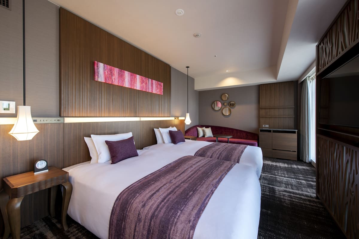 Quarto de hotel com duas camas, cabeceira de madeira, uma grande pintura abstrata, sofá vermelho e televisão.