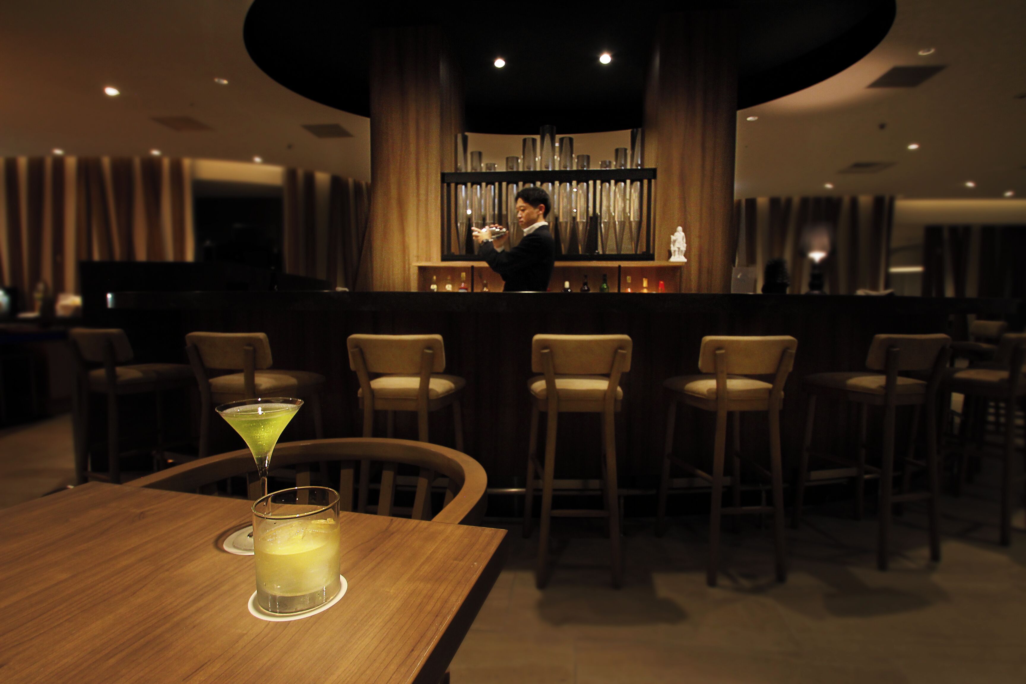 A bar area with a wooden table, bar stools, and a bartender.