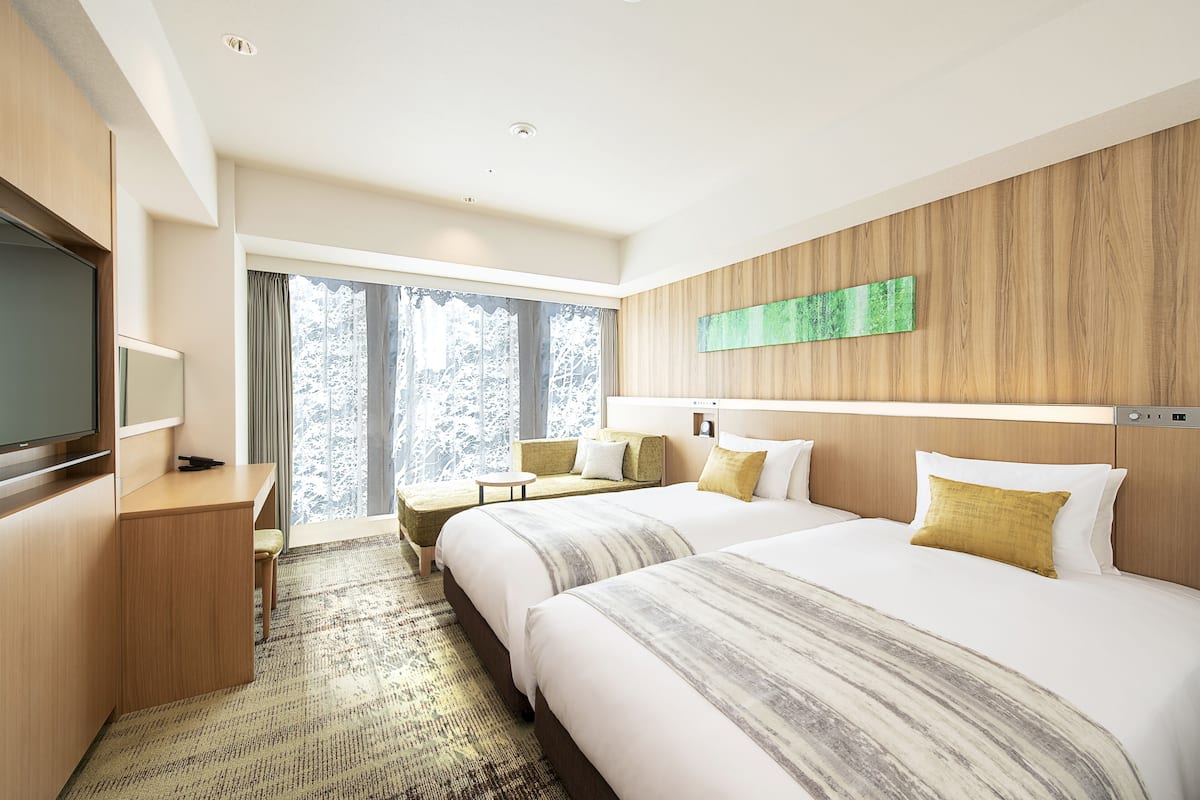 Quarto de hotel com duas camas, uma escrivaninha, televisão e uma janela grande com vista para árvores.