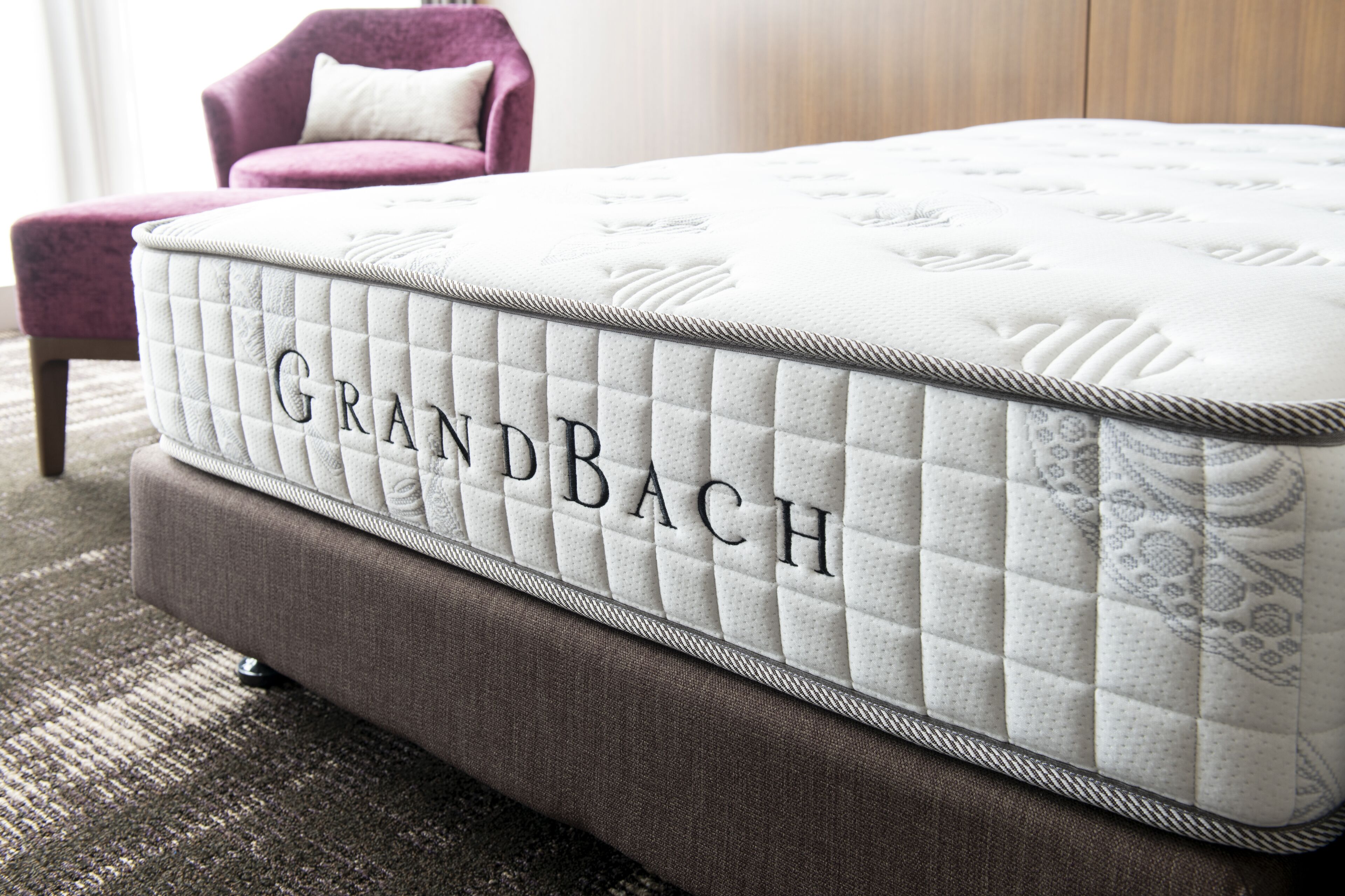 A Grandbach mattress with a quilted design, placed on a brown upholstered bench in a room with a patterned carpet.