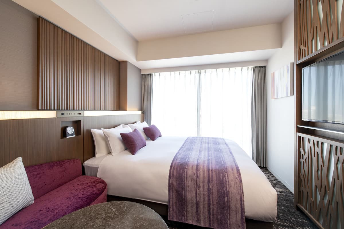 Quarto de hotel com uma cama grande, sofá, televisão e janela com cortinas.