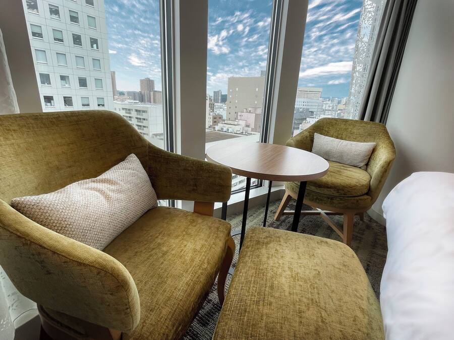 A hotel room with a large window, two armchairs, a small round table, and a bed.