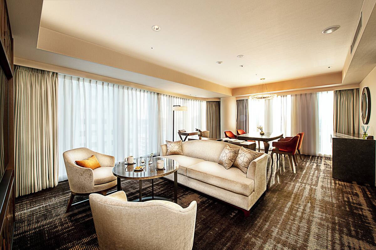 Quarto de hotel com sofá, poltronas, mesa de jantar e uma janela grande com cortinas.