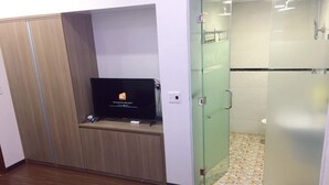 A room with a television on a wooden cabinet, a glass shower enclosure, and a tiled floor. - Fasy Hotel (Vinh Phuc)