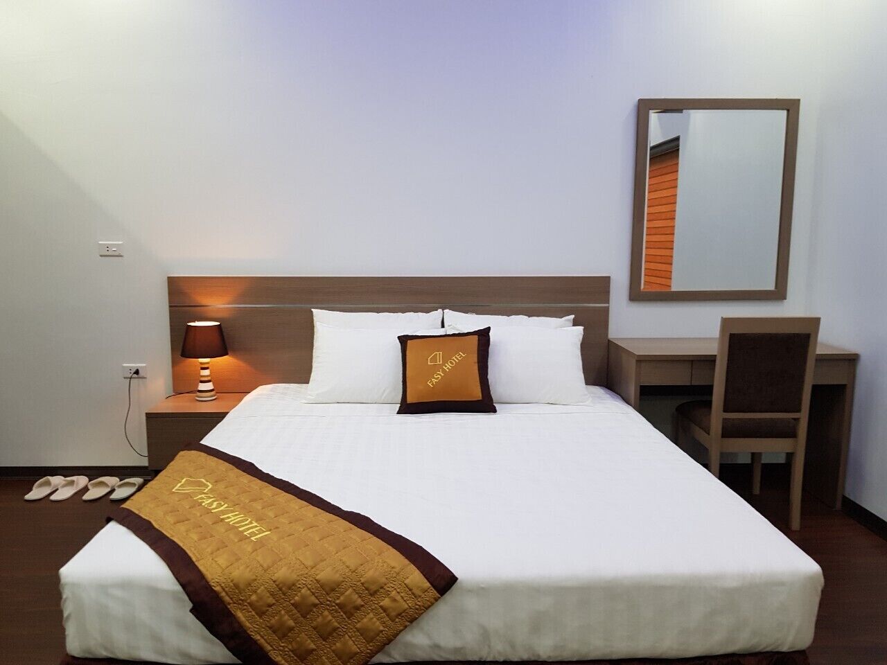 Una cama bien hecha con edredón blanco y manta marrón, una lámpara de noche, una mesita de madera, una silla y un espejo en la pared.