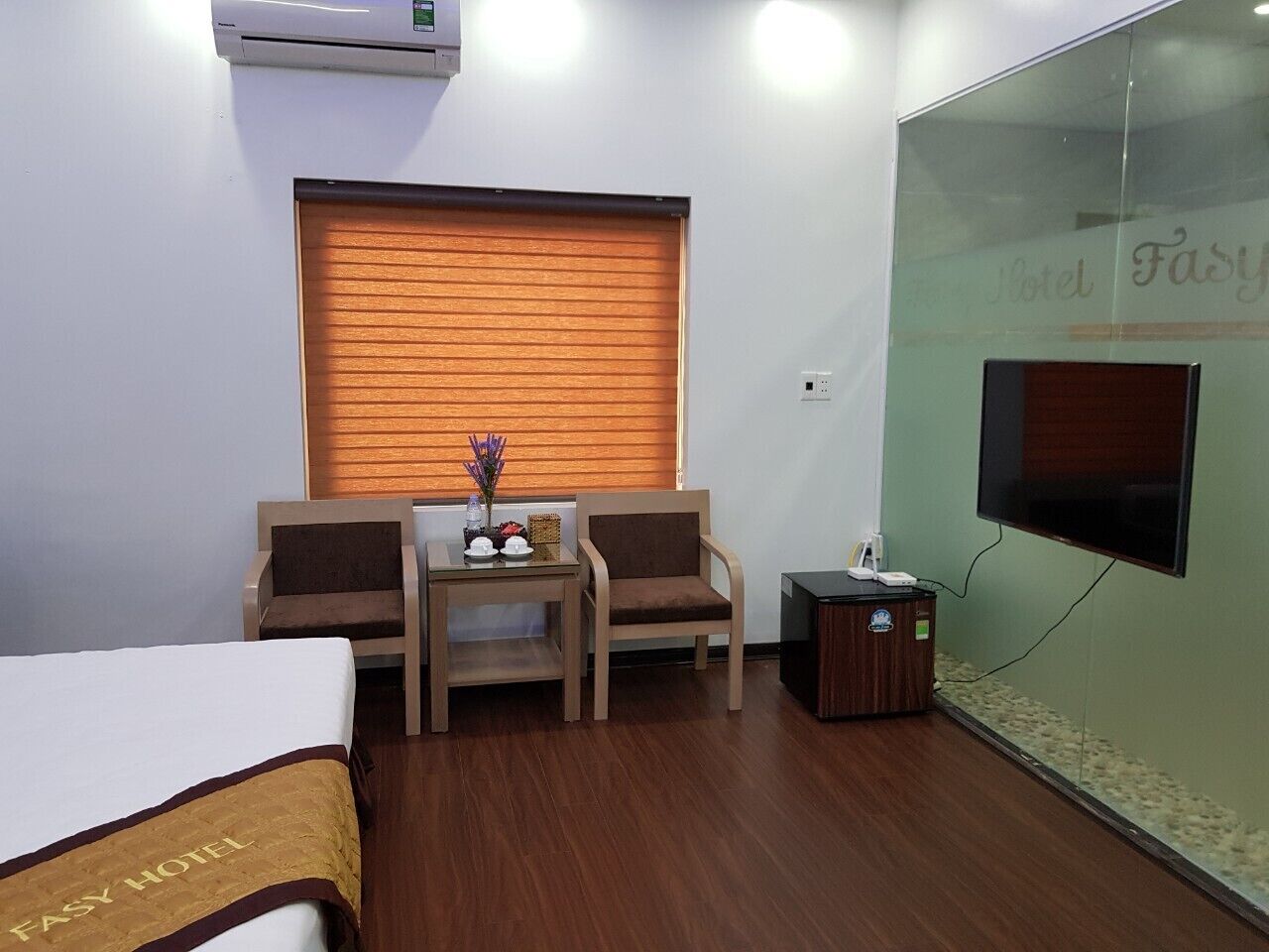 Habitación de hotel con una cama, dos sillas, una mesita con un florero, un televisor y un armario de madera.