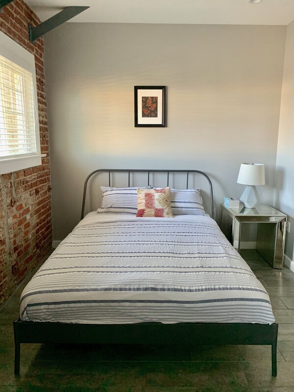 Una cama bien hecha con ropa de cama a rayas, una almohada decorativa y un cuadro en la pared, además de una mesita de noche con una lámpara.