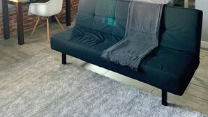 Una habitación con pared de ladrillo, cama con colcha a rayas, escritorio con lámpara y sofá negro.