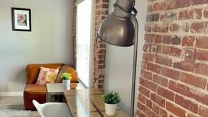 Habitación acogedora con pared de ladrillo, una mesa de madera, una silla blanca, un sofá de cuero marrón y una lámpara de pie.