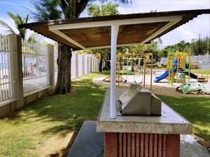 Un espace extérieur abrité avec un banc en béton, une aire de jeux avec des balançoires et des toboggans, et une zone délimitée par une clôture où se trouvent des palmiers.