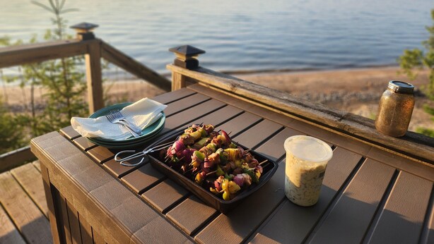 A wooden table with a plate of skewers, a jar of sauce, and a napkin on a deck overlooking the sea.