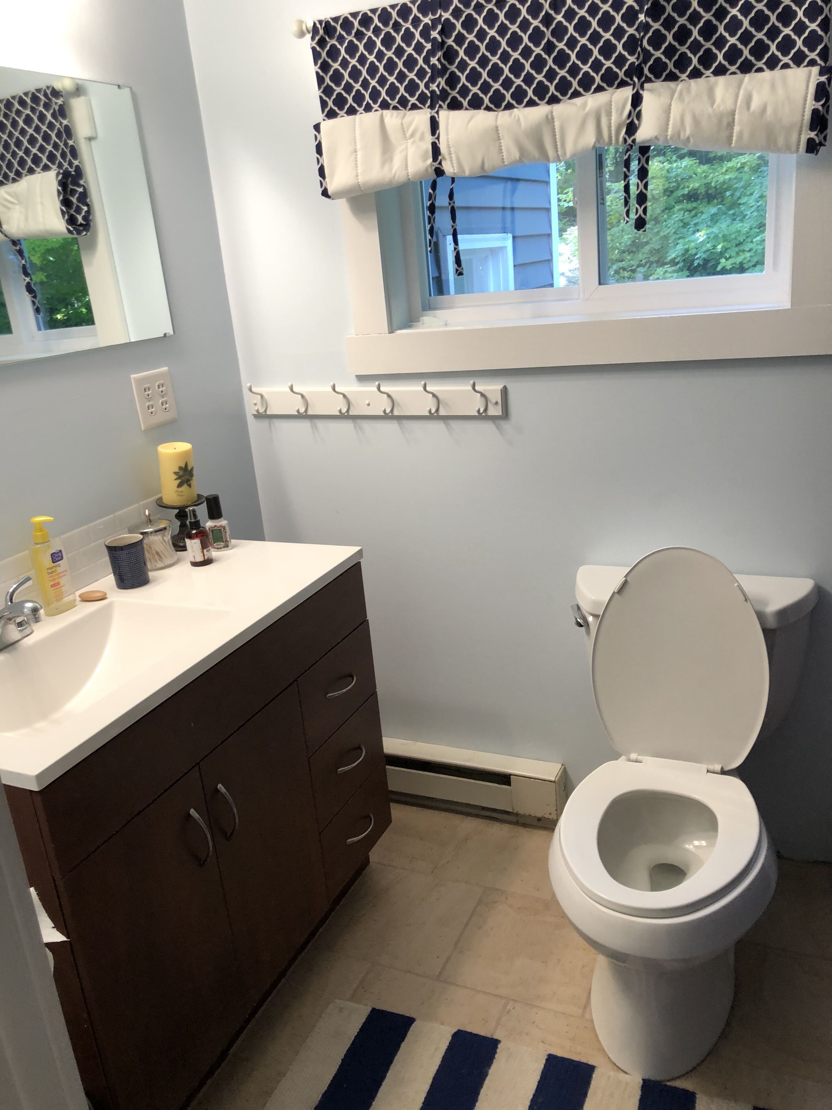 Une salle de bain avec un lavabo blanc, des meubles de rangement brun foncé, des toilettes et une fenêtre agrémentée d’une lambrequin à motifs.