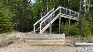 A wooden deck with metal railings leading to a staircase, surrounded by a pebbled area and trees.